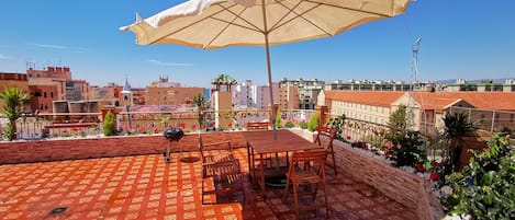 A rooftop terrace with a patterned red floor, a wooden table and chairs, a barbecue grill, and a large beige umbrella.