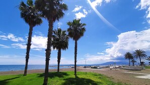 Una playa con palmeras, cielo despejado y barcos a lo lejos.