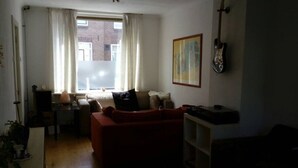 Ein Wohnzimmer mit einem roten Sofa, einem Couchtisch und einem Fenster mit Vorhängen. An der Wand hängt eine Gitarre und über dem Sofa hängt ein Bild.