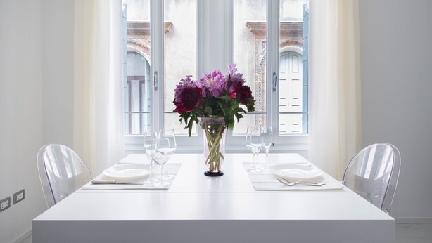 Mesa de jantar com um vaso de flores, cadeiras transparentes e vista para um edifício através de grandes janelas.