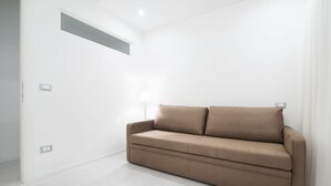 Quarto minimalista com sofá bege, piso claro e parede branca com uma pequena janela.