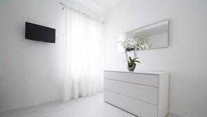 Quarto minimalista com cômoda branca, televisão de tela plana e uma orquídea em vaso.