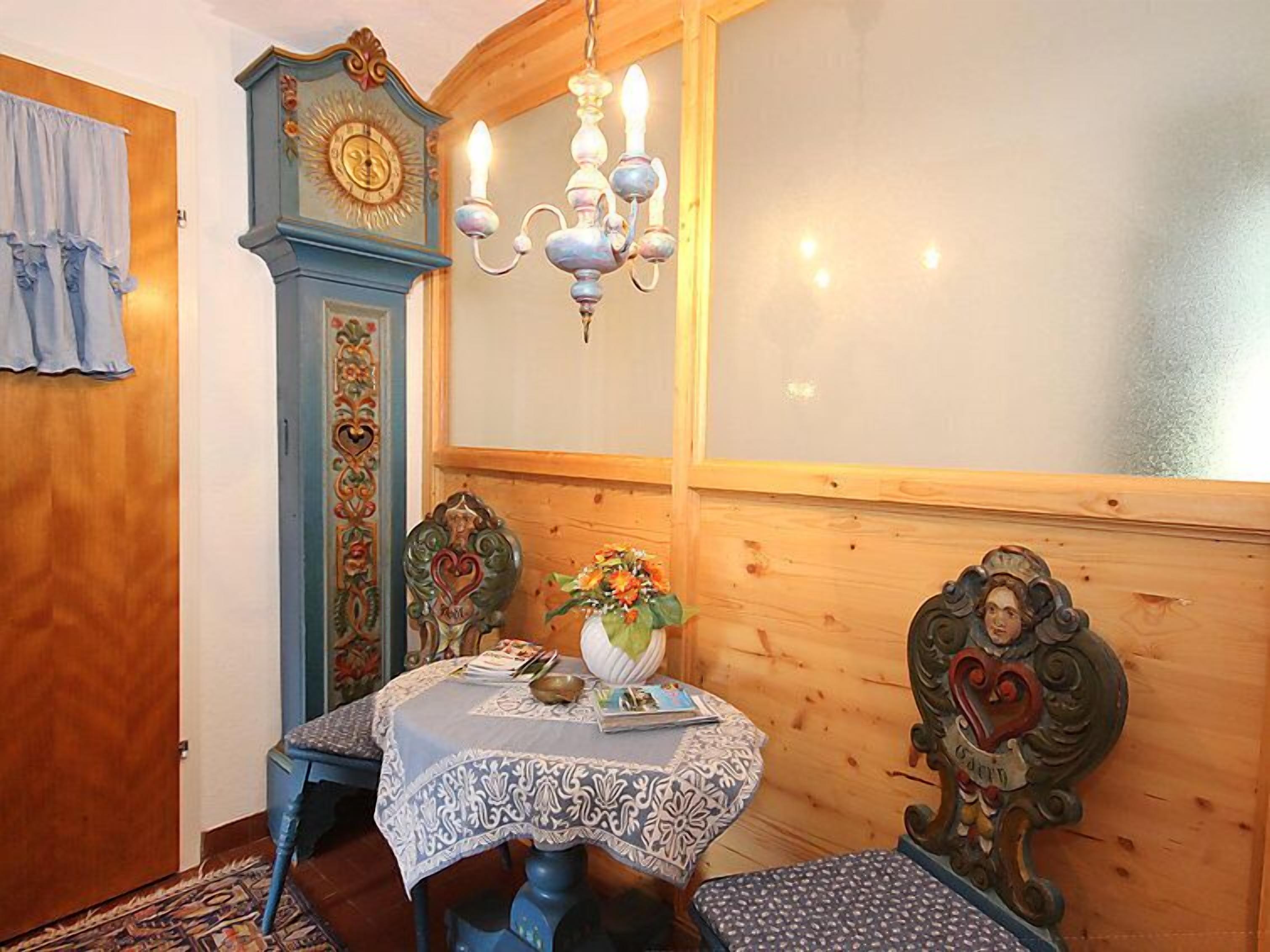 A room with a wooden wall, a blue cabinet with a clock, a chandelier, a table with a lace tablecloth, and a bench.