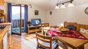 A cozy kitchen with a dining area, wooden furniture, a TV, and a balcony with curtains.