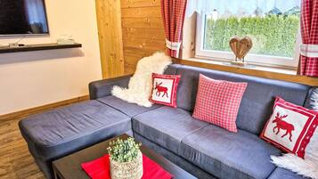 Ein modernes Wohnzimmer mit einem grauen Sofa, roten und weißen Kissen in Rautenmuster, einem Holz-TV-Schrank und einem Fenster mit Blick auf Grünflächen.