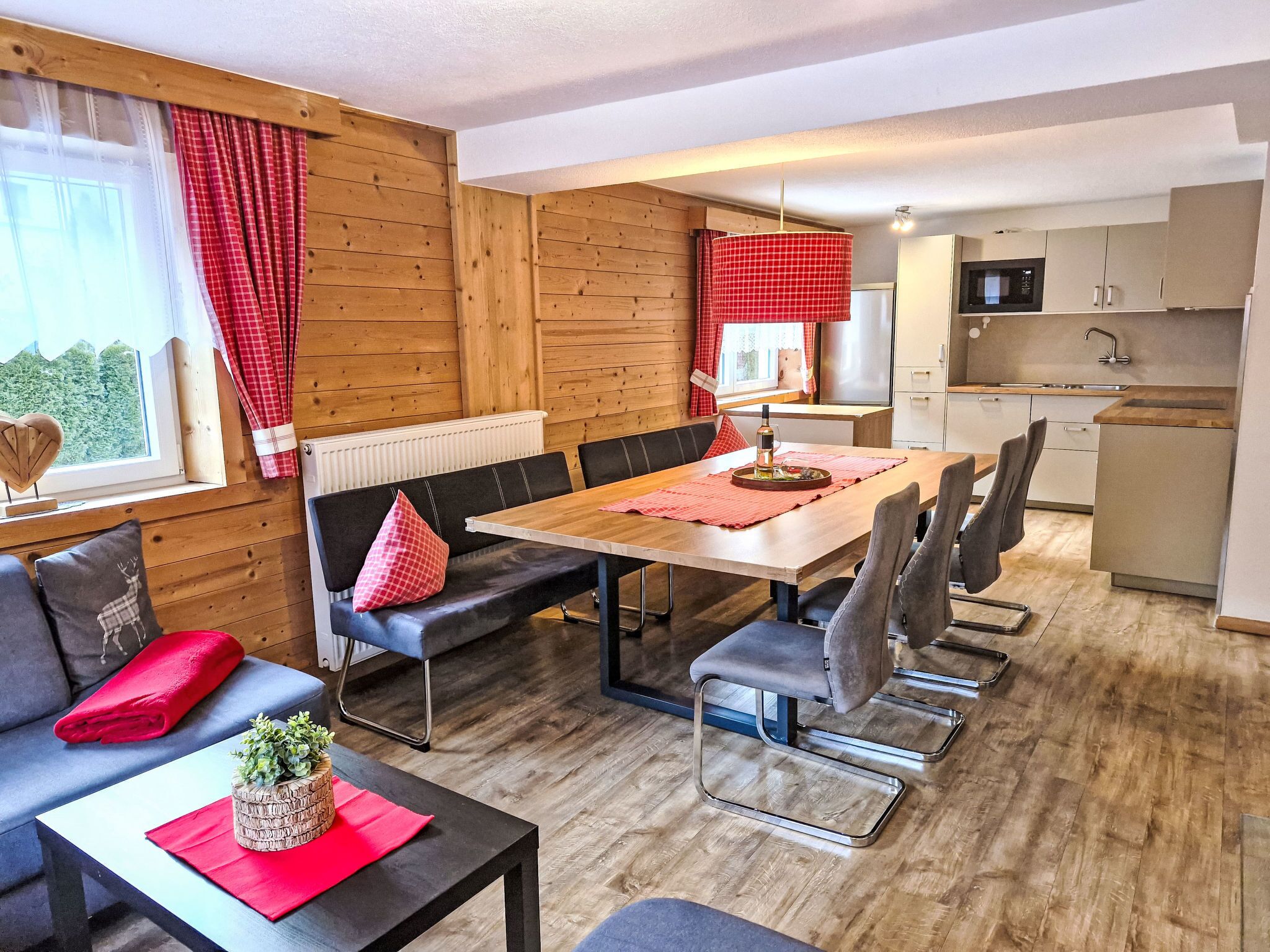 Ein moderner, hell eingerichteter Raum mit Holzboden, einem Esstisch mit Stühlen, einer Couch mit Kissen, einer kleinen Küchenzeile mit Mikrowelle und einem Fenster mit roten Vorhängen.