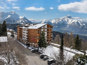 Un hotel de varios pisos con balcones, un centro de esquí al fondo y un paisaje cubierto de nieve.
