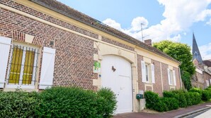 Un edificio de ladrillo con una puerta blanca, un pequeño jardín y la aguja de una iglesia al fondo.