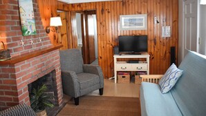 A cozy living room with a brick fireplace, a television on a wooden stand, a blue sofa, and a checkered armchair.