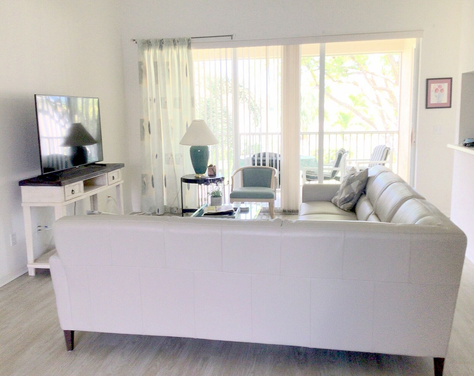 A living room with a white sofa, a television on a stand, a lamp, and a view of the outdoors through large windows.