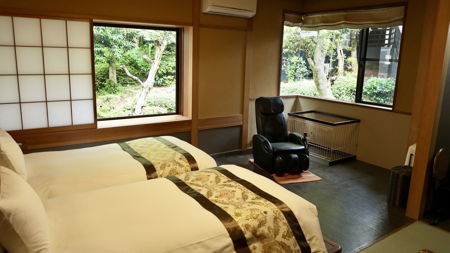 Habitación con dos camas, una silla de masaje, una jaula pequeña para mascotas y ventanas grandes con vistas a zonas verdes.
