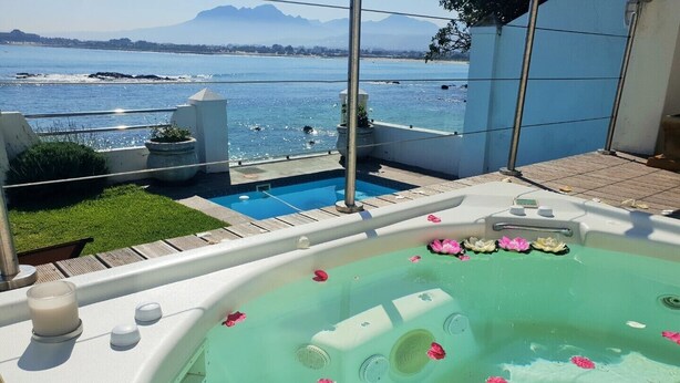 A hot tub with pink flower petals floating on the water, surrounded by a pool and a view of the sea and mountains. - Westbank (Cape Town)