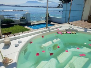A hot tub with pink flower petals floating on the water, surrounded by a pool and a view of the sea and mountains. - Westbank (Cape Town)