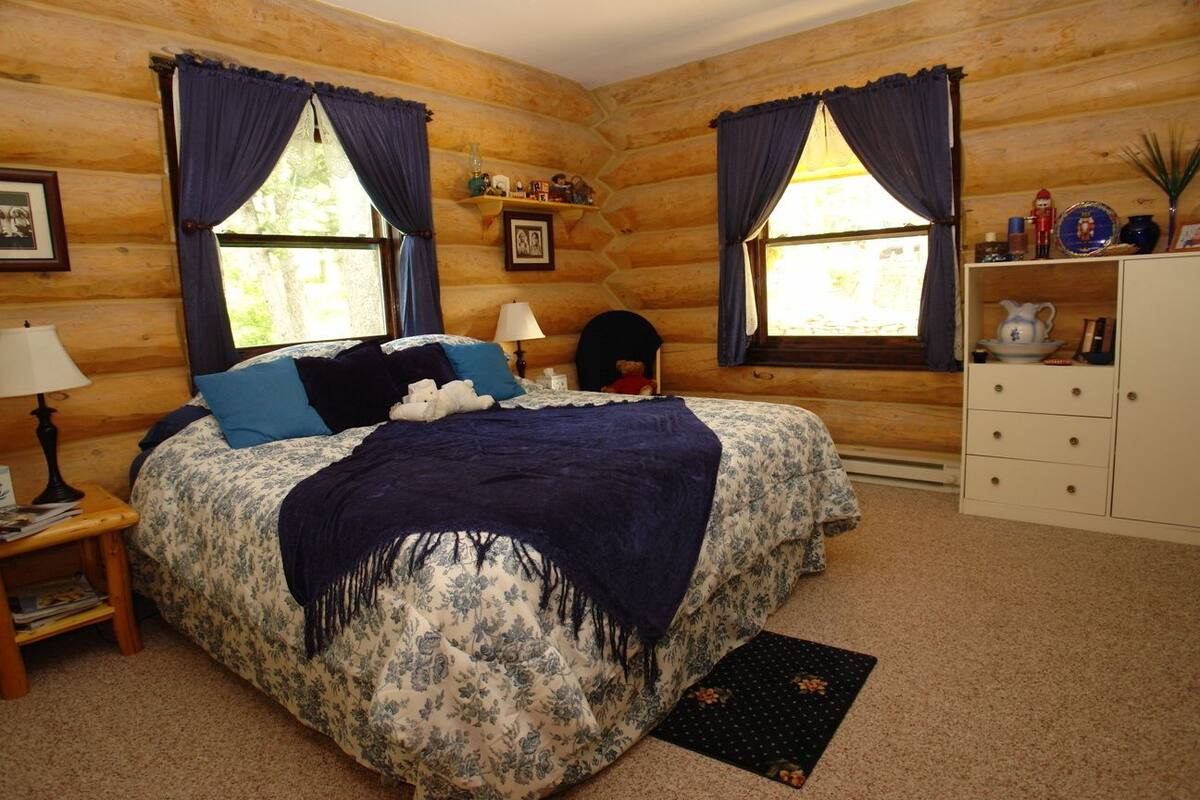 A log cabin bedroom with a bed, two windows with curtains, a nightstand, a dresser, and a small shelf with decorative items.