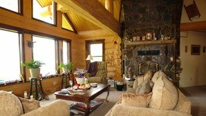 A cozy log cabin interior with a stone fireplace, wooden beams, and large windows. - The Garrison Inn a Montana Bed & Breakfast (Kalispell)