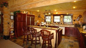 A log cabin kitchen with a central island, wooden bar stools, and a stone wall. - The Garrison Inn a Montana Bed & Breakfast (Kalispell)