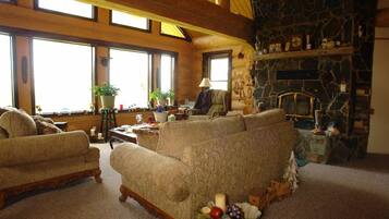 A cozy living room with a stone fireplace, large windows, and a mix of furniture including sofas and a coffee table.