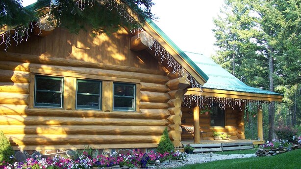 A log cabin with a stone foundation, a porch with a bench, and a flower bed with various flowers. - The Garrison Inn a Montana Bed & Breakfast (Kalispell)