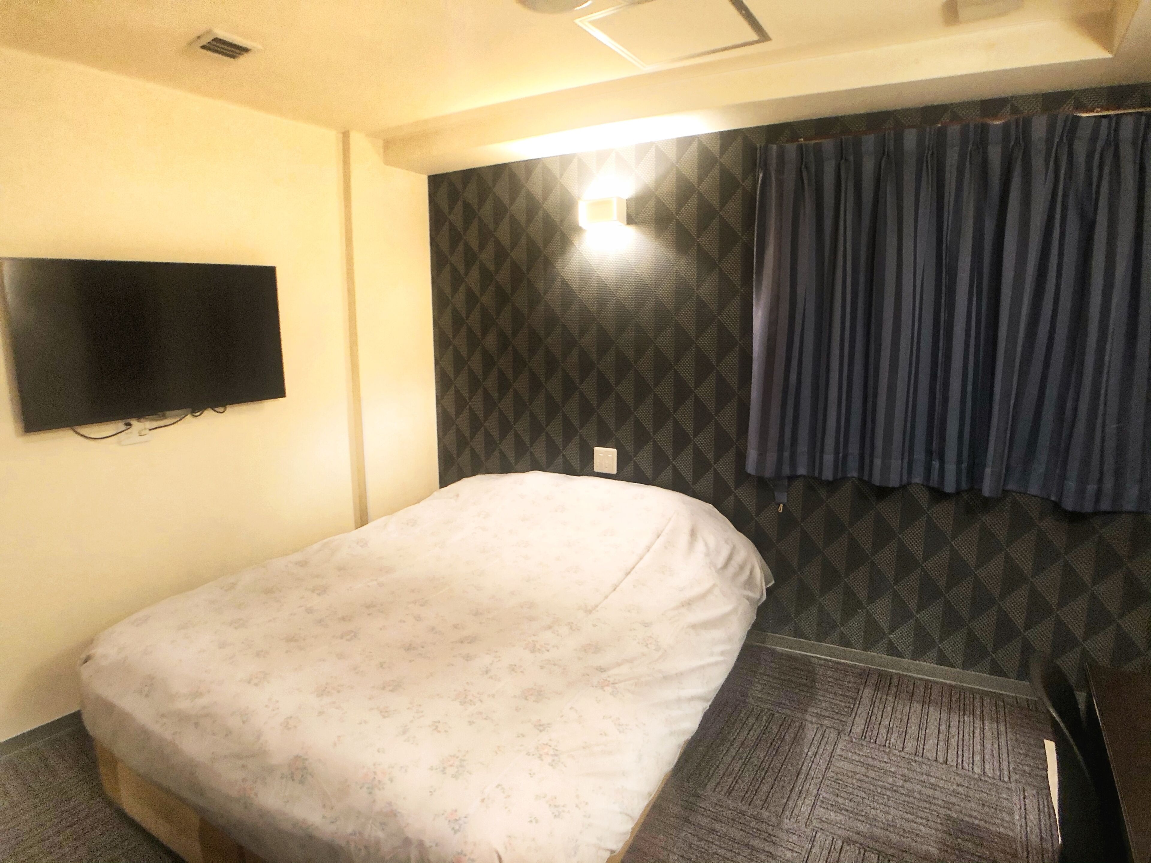 A small, single-bed room with a patterned bedspread, a mounted television, and a patterned wall.