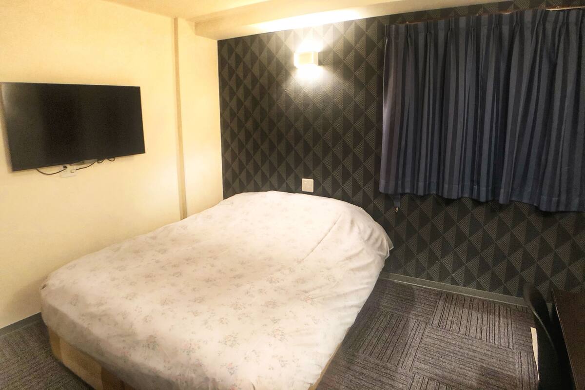 Quarto pequeno com uma cama de solteiro, colcha estampada, televisão na parede e decoração padronizada.
