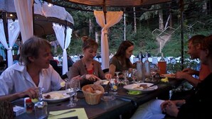 Un grupo de personas cenando en una mesa al aire libre bajo una cubierta, rodeado de paisajes naturales.