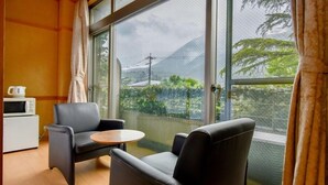 A room with two black leather chairs, a small round table, a white microwave, and a white refrigerator. There is a large window with a view of greenery and a power line. - Yufuin Floral Village Hotel (Yufu)