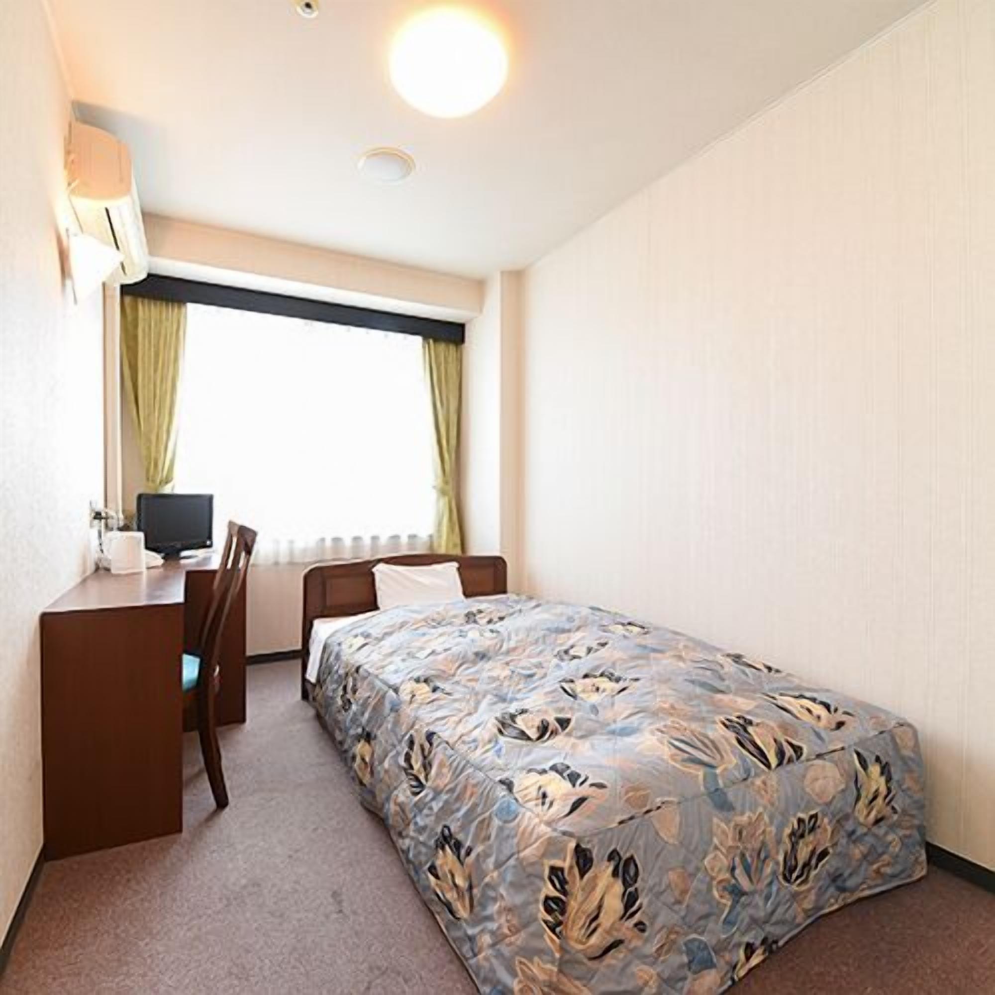 Ein Zimmer mit einem Bett, einem Schreibtisch, einem Stuhl, einem Fenster mit Vorhängen und einer Wandlampe.