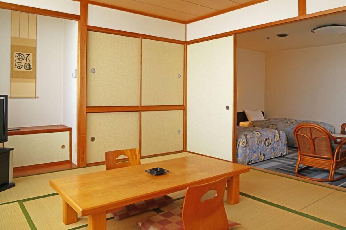 Une pièce de style japonais traditionnel, avec un sol recouvert de tatamis, une table basse, des chaises en bois, un lit recouvert d’une courtepointe à motifs floraux bleus et un téléviseur posé sur un meuble.