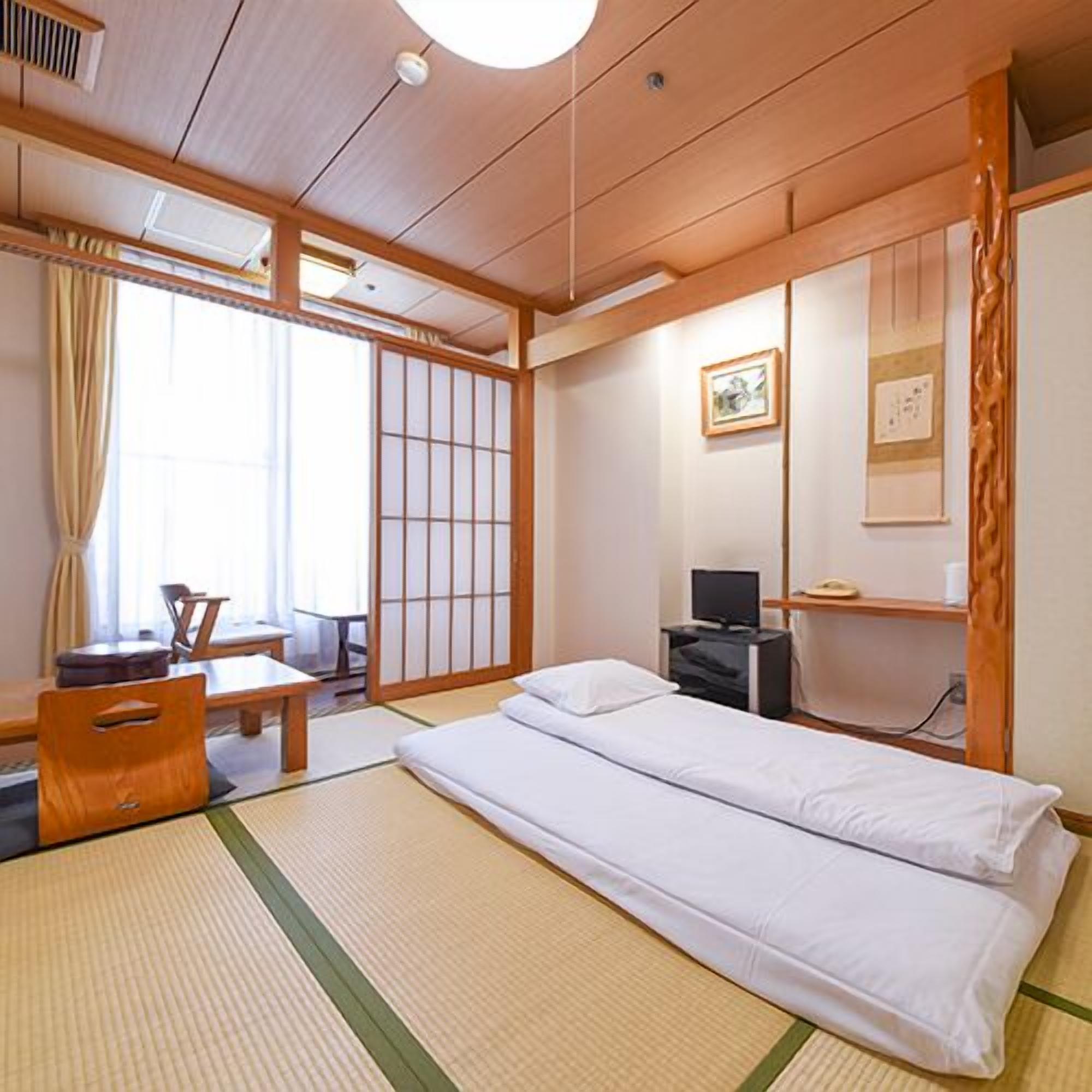 Ein traditioneller japanischer Raum mit Tatami-Bodenbelag, niedrigem Bett, Schreibtisch mit Stuhl und verschiebbaren Shoji-Schiebetüren.