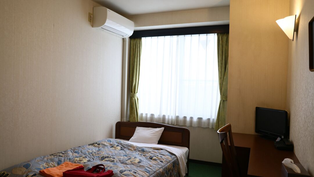 A hotel room with a bed, a desk, a chair, a television, and an air conditioning unit.