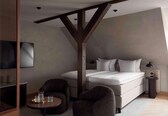 A modern hotel room with a bed, two armchairs, a small table with glasses, a TV, and a wooden ceiling structure.