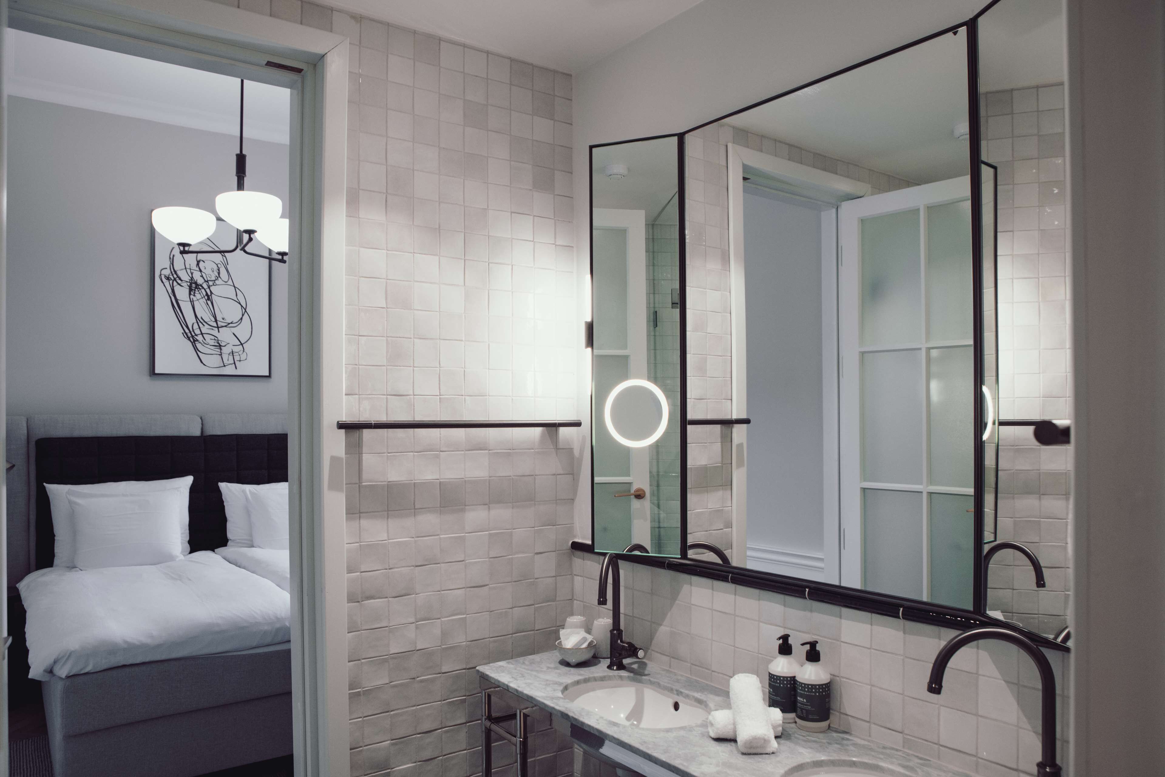 A modern bathroom with a large mirror, a sink with toiletries, and a bed with white linens.