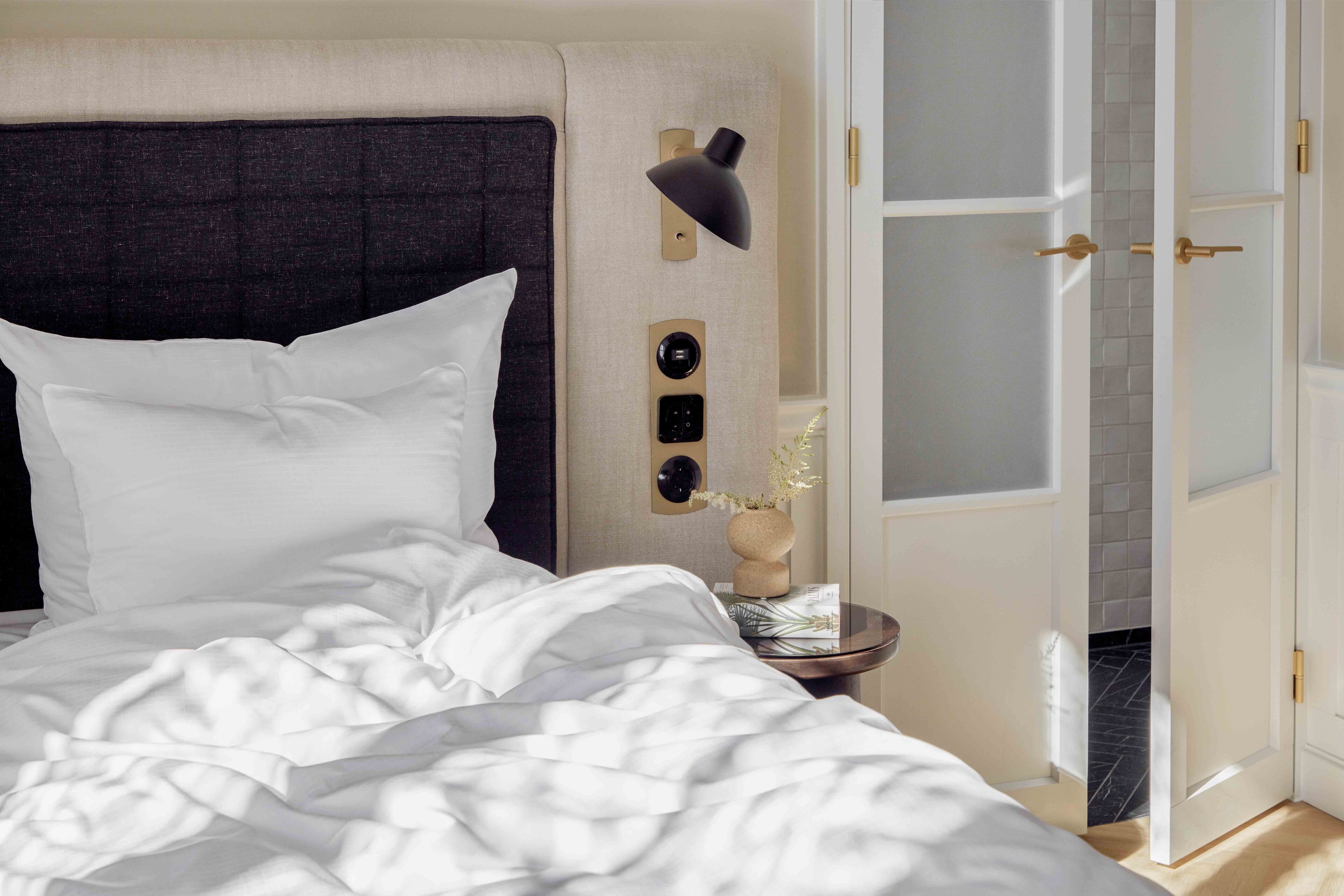 Una cama bien tendida con sábanas blancas, cabecera oscura, mesita de noche con lámpara y puerta que da a un baño con pared de azulejos.