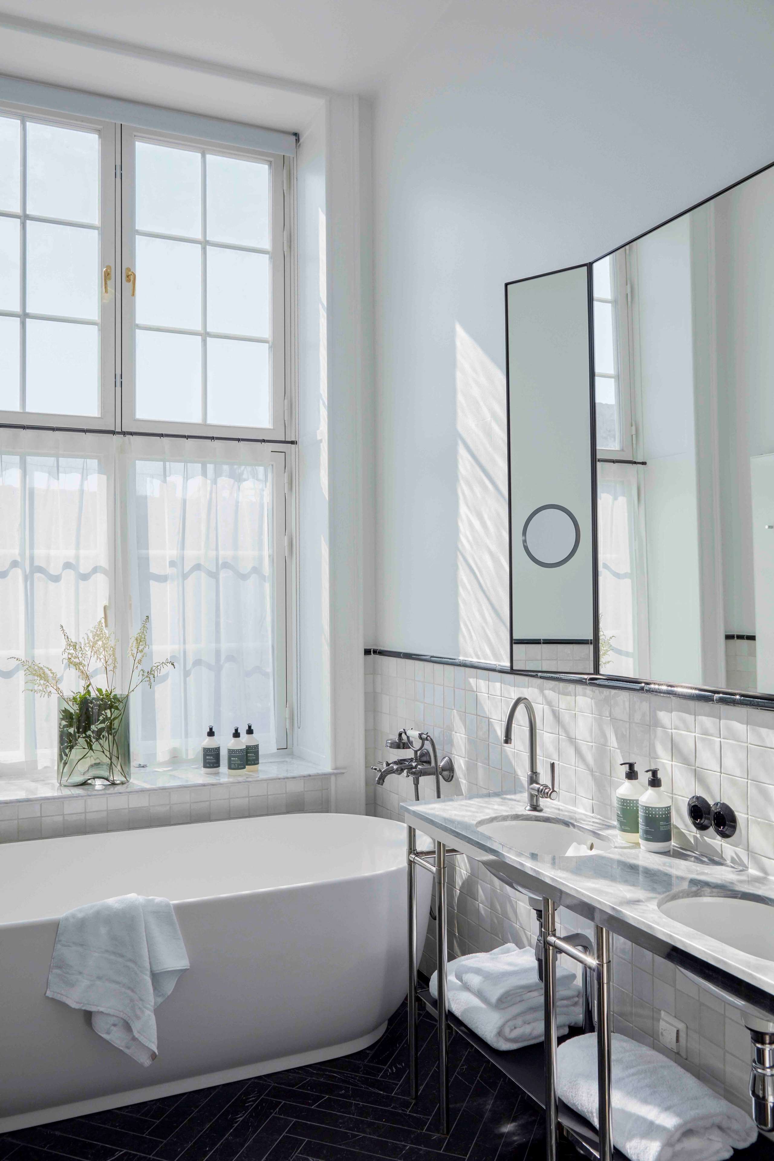Un baño moderno con una bañera independiente, un espejo grande y una ventana con cortinas de gasa.