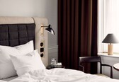 A hotel room with a bed, a bedside table with a lamp, a chair, and a window with curtains.