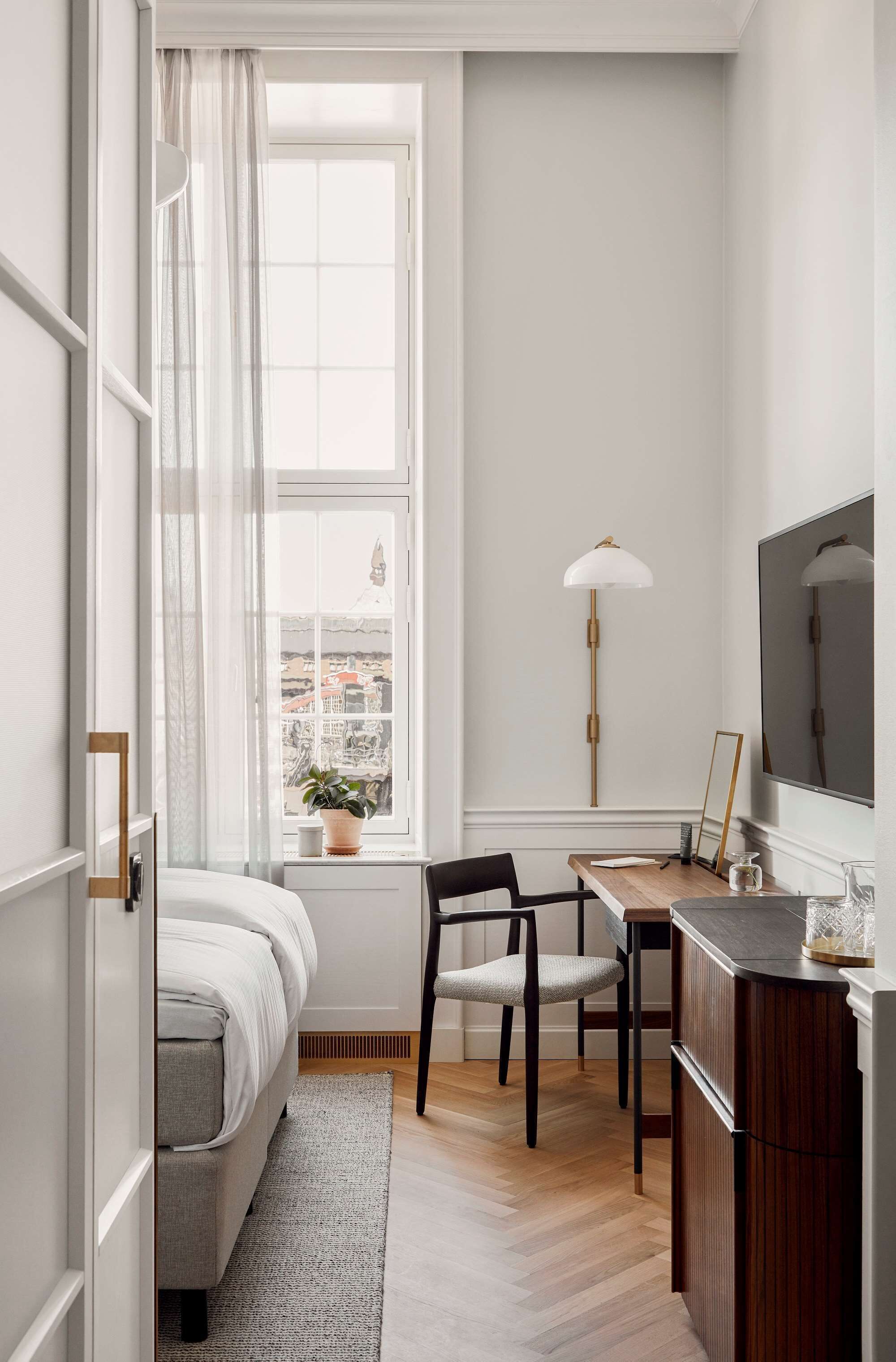 Een kleine slaapkamer met een bed, nachtkastje, stoel en een raam met gordijnen.