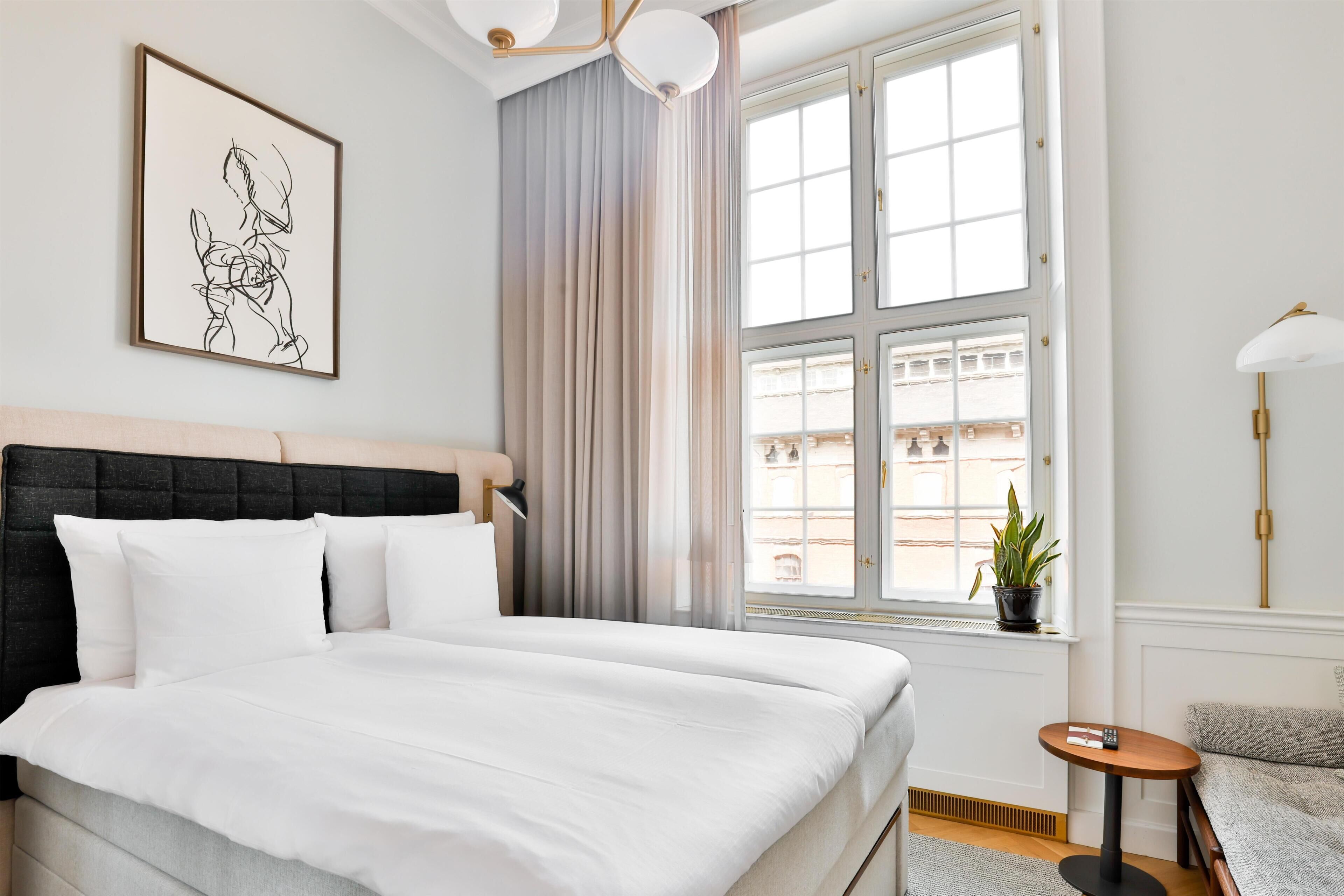 Una cama bien hecha con sábanas blancas, un cabecero negro, una obra de arte enmarcada en la pared, una ventana con cortinas y una pequeña mesa redonda con una lámpara.