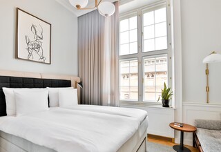 A neatly made bed with white linens, a black headboard, a framed artwork on the wall, a window with curtains, a small round table, and a lamp.