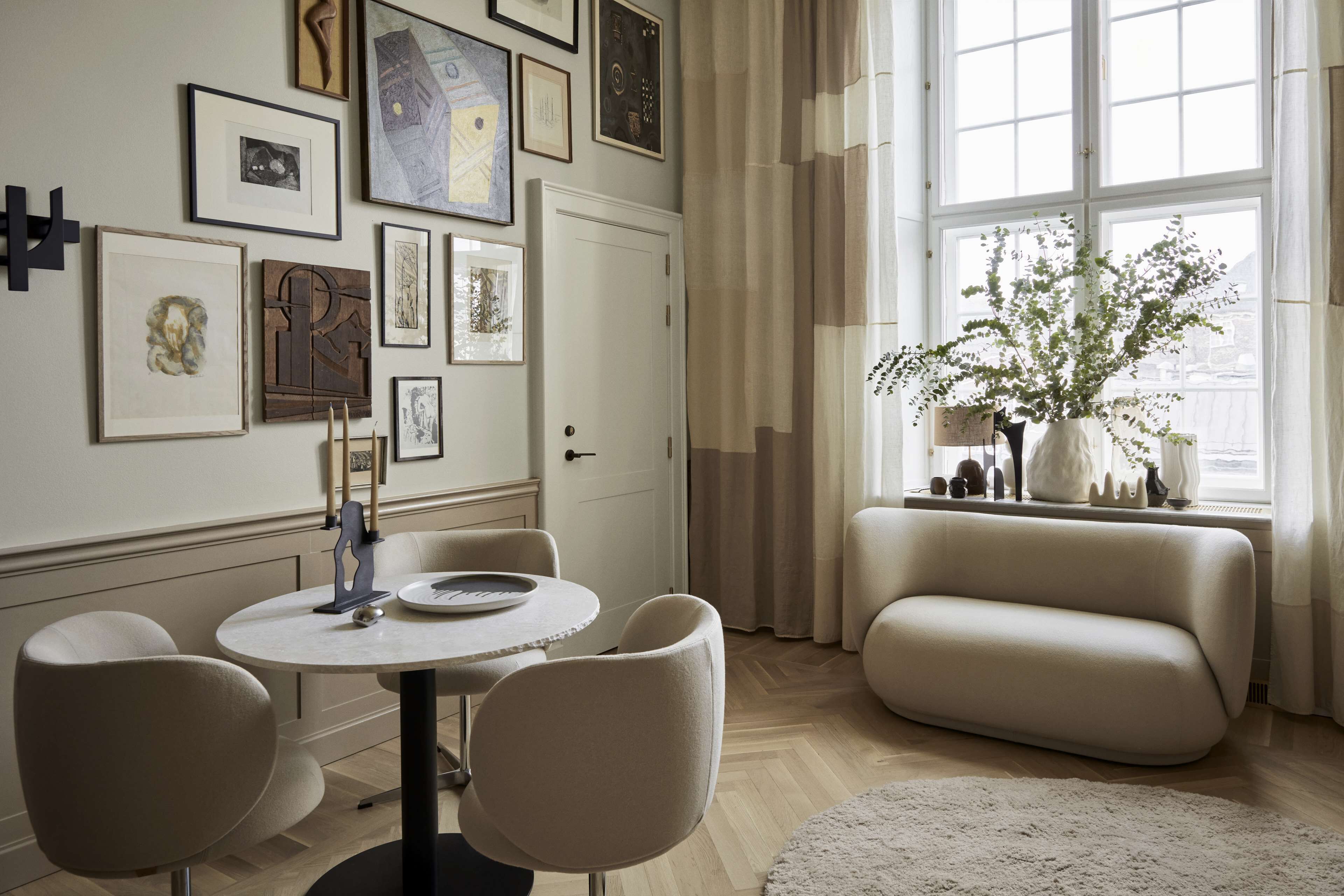 A modern living room with a round table, two chairs, a sofa, and a large window with curtains.