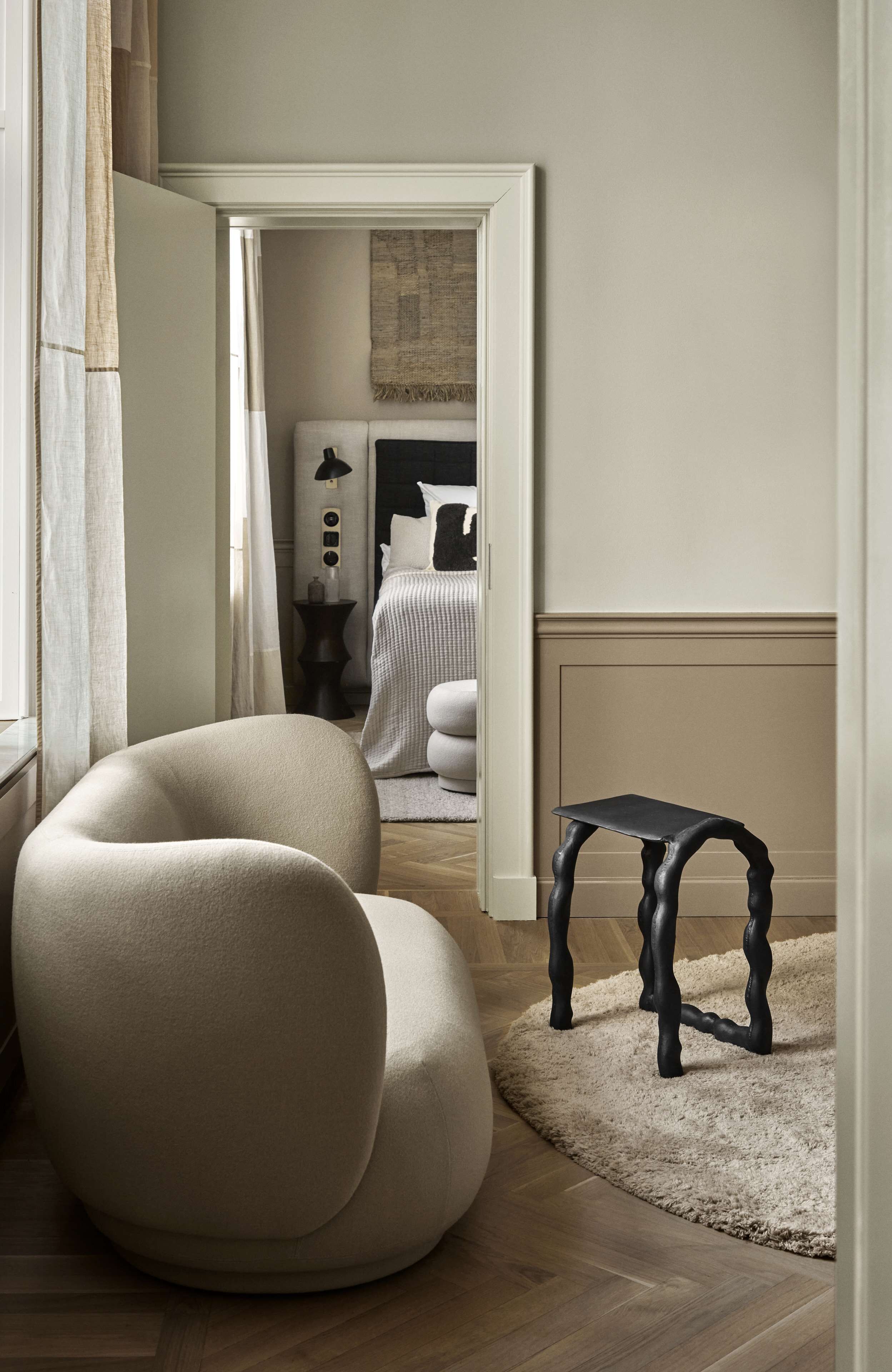 A modern living room with a curved sofa, a small black table, and a view into another room with a bed and a nightstand.