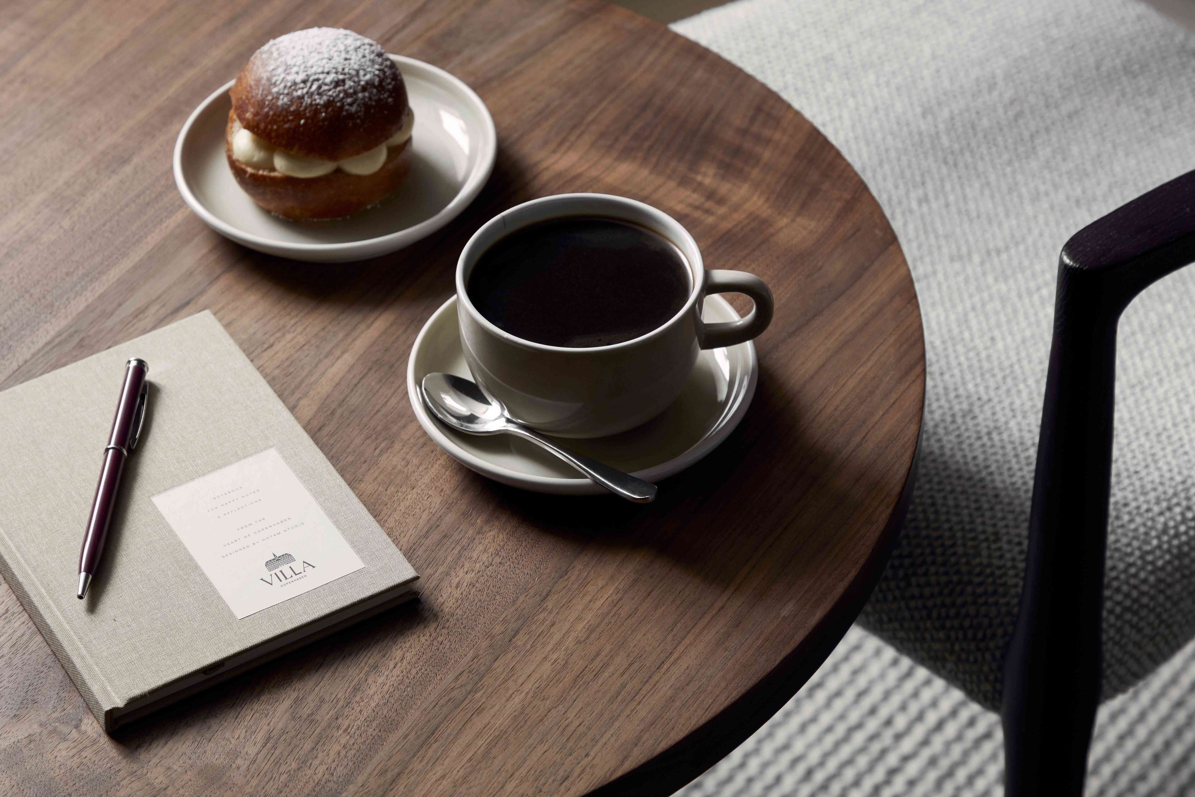 A wooden table with a cup of coffee, a pastry, a notebook, and a pen.