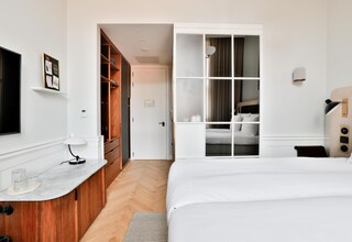 A hotel room with a bed, a wooden wardrobe, a desk with a lamp, and a television on the wall.
