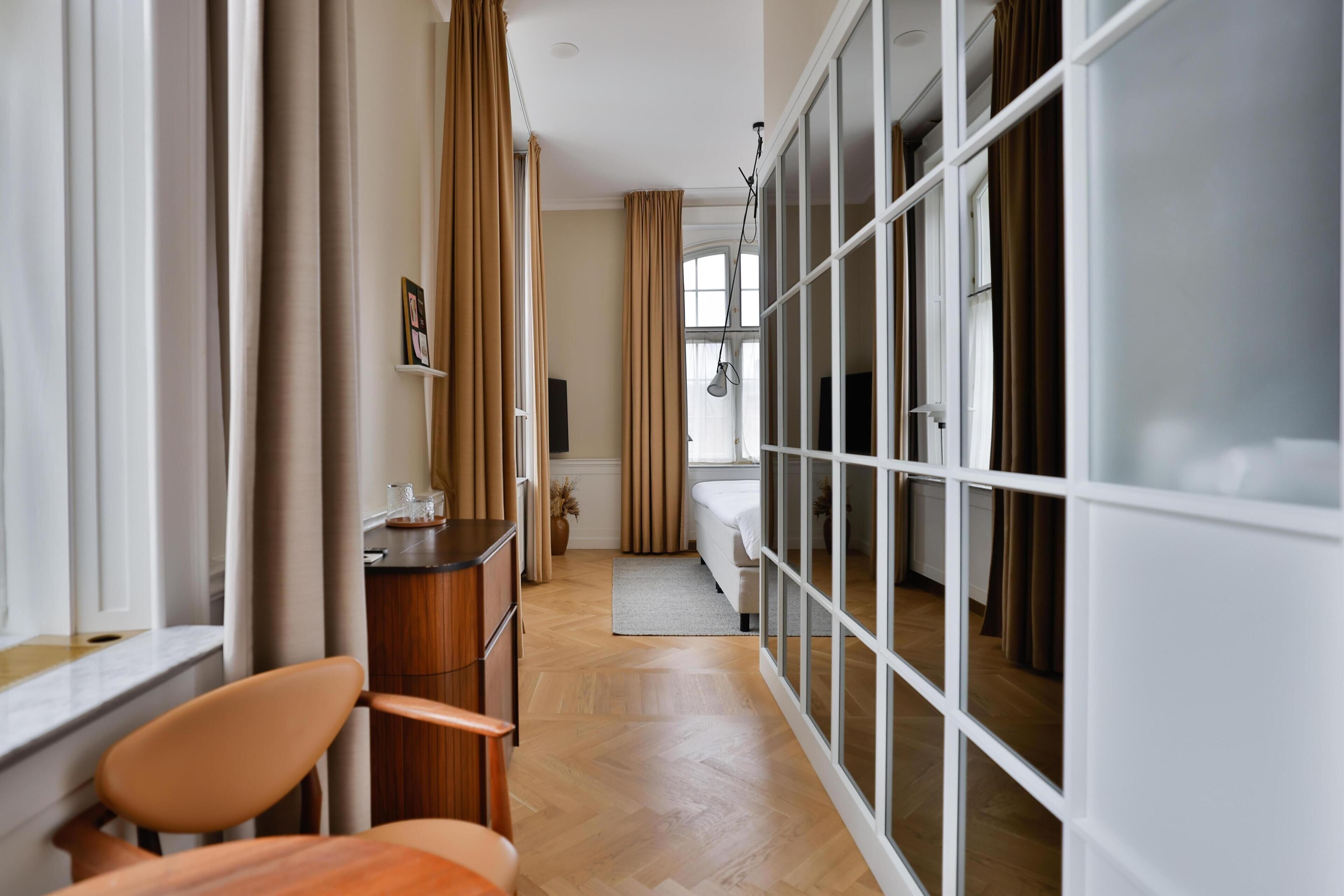 A hotel room with a wooden floor, a desk with a chair, a bed, and a large window with curtains.