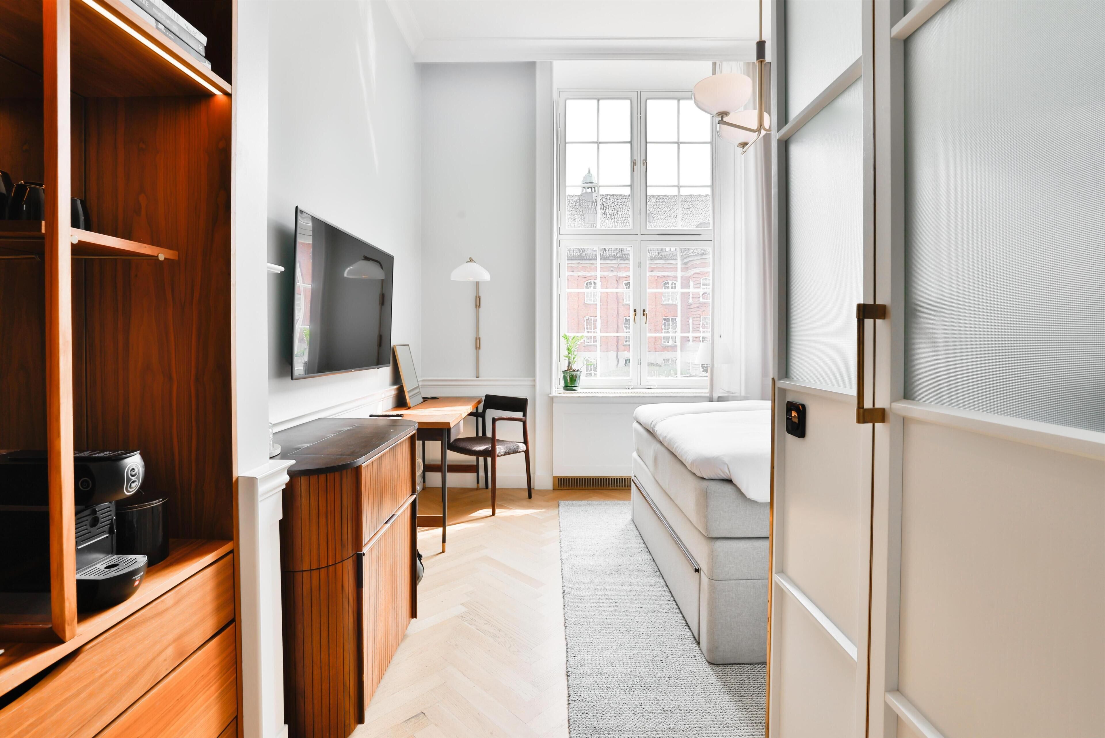 Habitación pequeña con cama, escritorio, televisión y ventana con vistas a edificios.