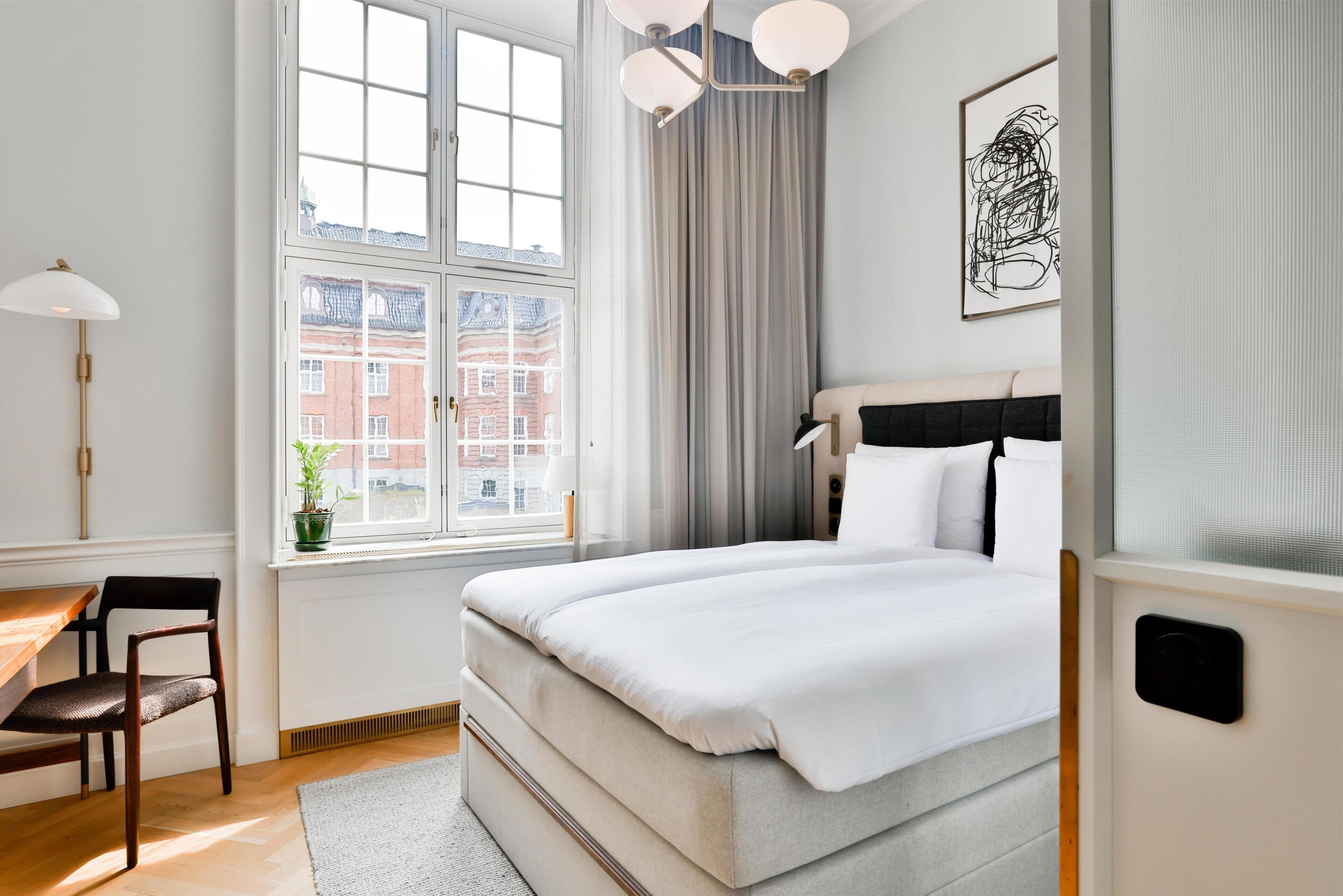 Un dormitorio con cama, escritorio, silla, ventana con cortinas y vista a edificios.