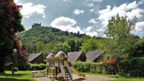 A playground with a slide and climbing structures, surrounded by a grassy area with a castle in the background. - Camping le Paisserou - Chalets (Najac)