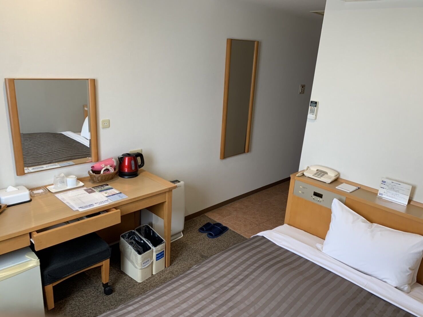 A hotel room with a bed, a desk with a telephone, a mirror, a radiator, and a trash bin.