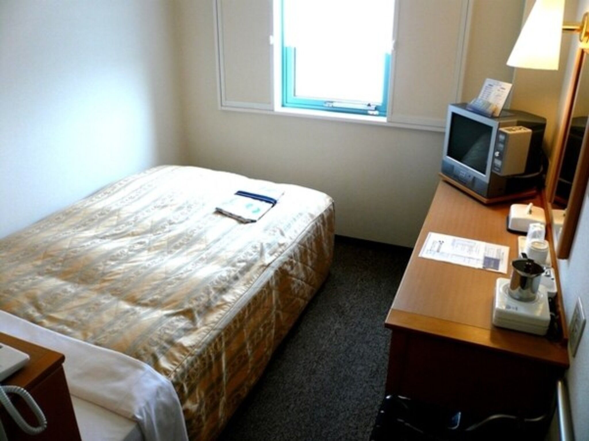 A small hotel room with a single bed, a desk with a computer and a lamp, and a window with blinds.