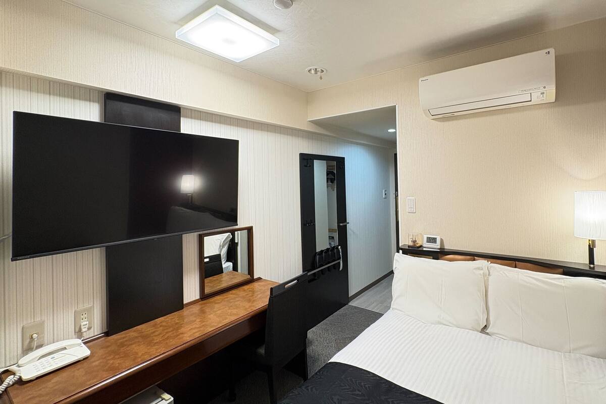 Habitación de hotel con cama, escritorio con teléfono, televisor de pantalla plana, espejo, armario y equipo de aire acondicionado.