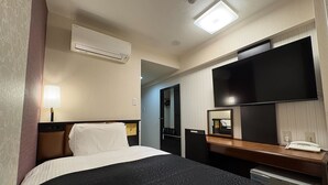 Quarto de hotel com cama, luminária de cabeceira, ar condicionado, televisão, espelho e escrivaninha com telefone.